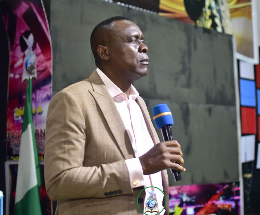 Apostle Prof. E. C. Ejiofor in a beige plaid blazer over a pale pink shirt, speaking into a blue microphone on a church stage with a Praiseland logo and Nigerian flag visible behind him