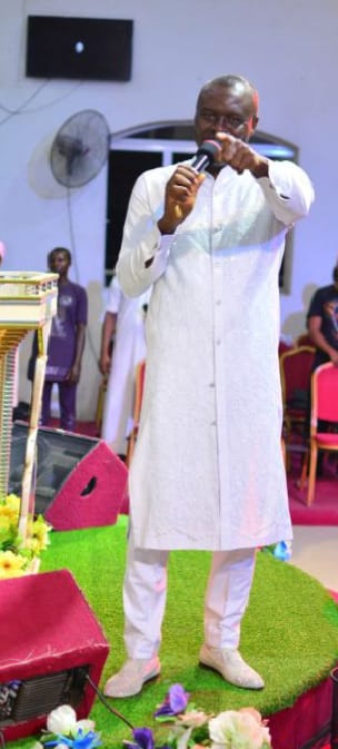 Apostle Prof. E. C. Ejiofor in a white robe, microphone in hand, pointing toward the congregation