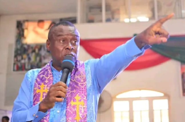 Apostle Prof. E. C. Ejiofor in a light blue robe and purple stole with gold crosses, preaching and pointing upward