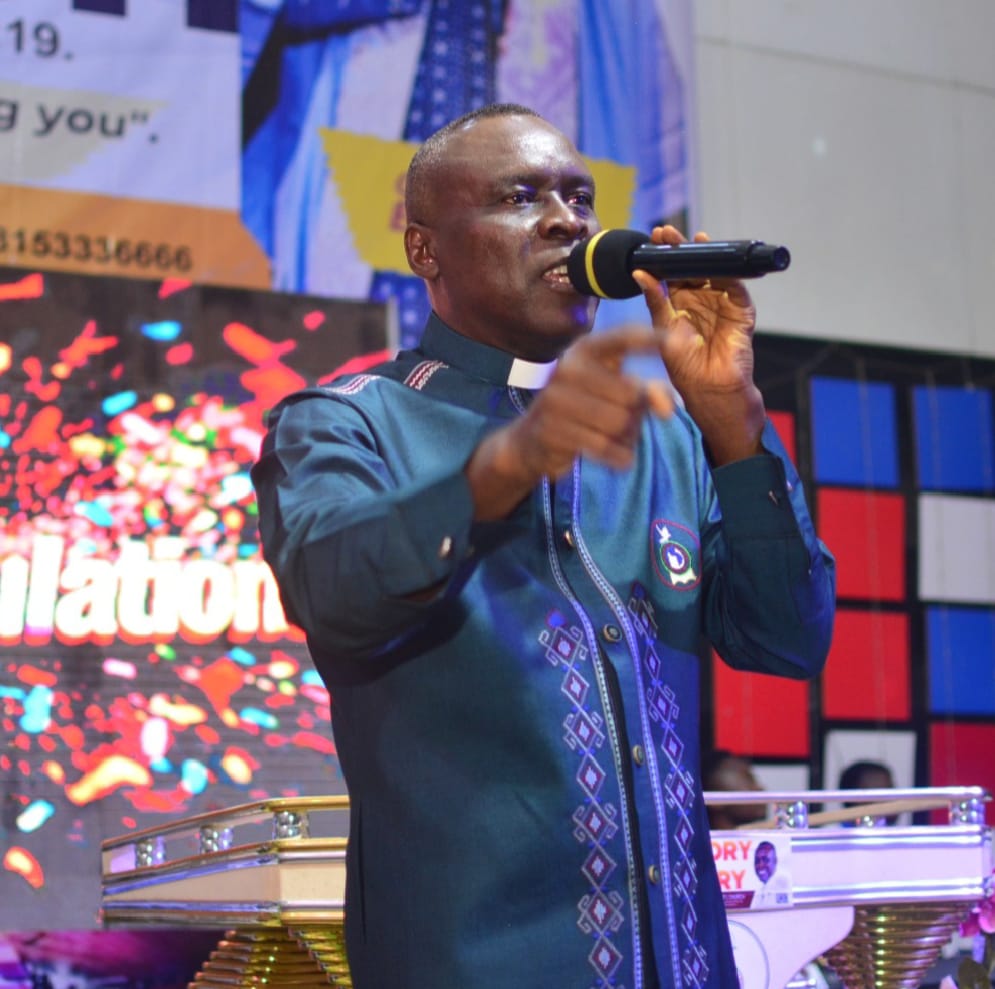 Apostle Prof. E. C. Ejiofor in a teal clerical shirt with diamond embroidery, pointing toward the congregation while preaching at a Praiseland Royal Springs Int'l Church service