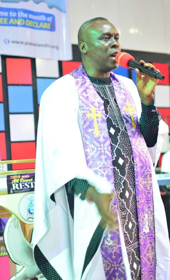 Apostle Prof. E. C. Ejiofor preaching into a microphone with a red windscreen, wearing a voluminous white robe and a purple stole embroidered with gold crosses and a black-and-white diamond pattern, framed by a red, blue and white panelled wall