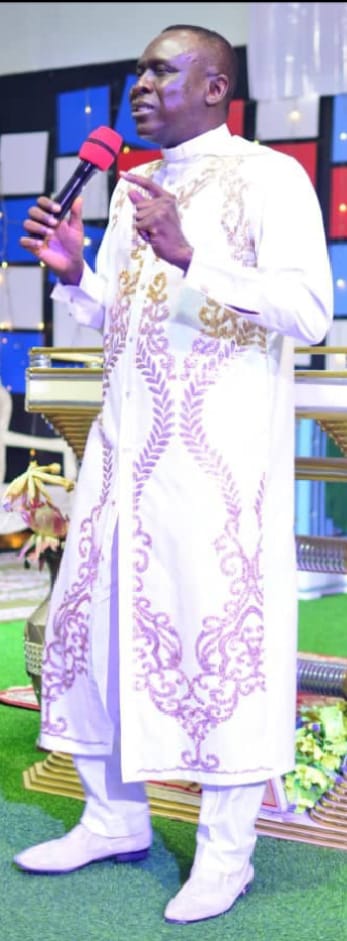 Apostle Prof. E. C. Ejiofor preaching on a stage in a white clerical robe with gold and purple embroidery, holding a microphone and gesturing — General Overseer of Praiseland Royal Springs Int'l Church