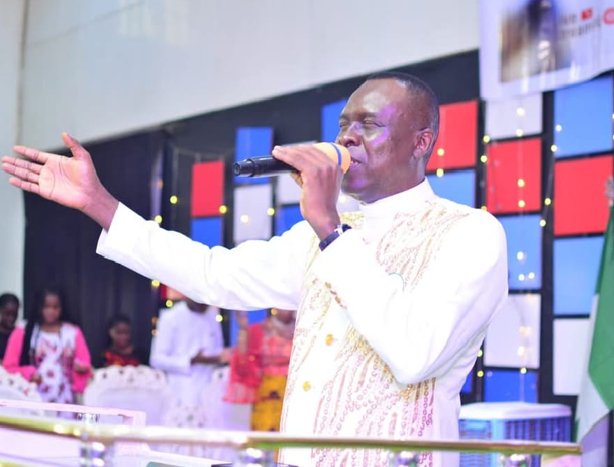Apostle Prof. E. C. Ejiofor preaching with a microphone, wearing a white robe with gold embroidery, one arm extended