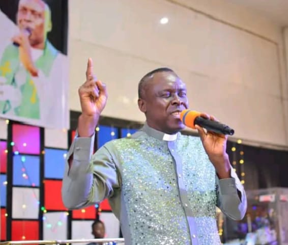 Apostle Prof. E. C. Ejiofor preaching in a light green sequined robe with a clerical collar, holding a microphone with one finger raised at Praiseland Royal Springs Int'l Church