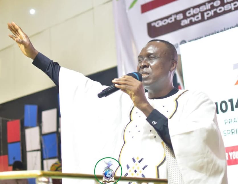 Apostle Prof. E. C. Ejiofor in a white embroidered robe, preaching into a microphone on a church stage with a colorful backdrop