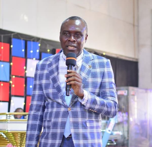 Apostle Prof. E. C. Ejiofor in a blue plaid blazer over a white shirt and light blue tie, holding a microphone with an orange windscreen on stage — General Overseer of Praiseland Royal Springs Int'l Church