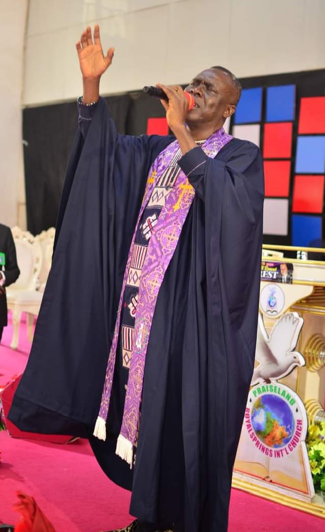 Apostle Prof. E. C. Ejiofor in a dark clerical robe with a purple embroidered stole, singing passionately into a microphone with his hand raised on stage — General Overseer of Praiseland Royal Springs Int'l Church