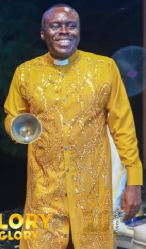 Apostle Prof. E. C. Ejiofor in a mustard-yellow embroidered clerical robe, holding a handbell