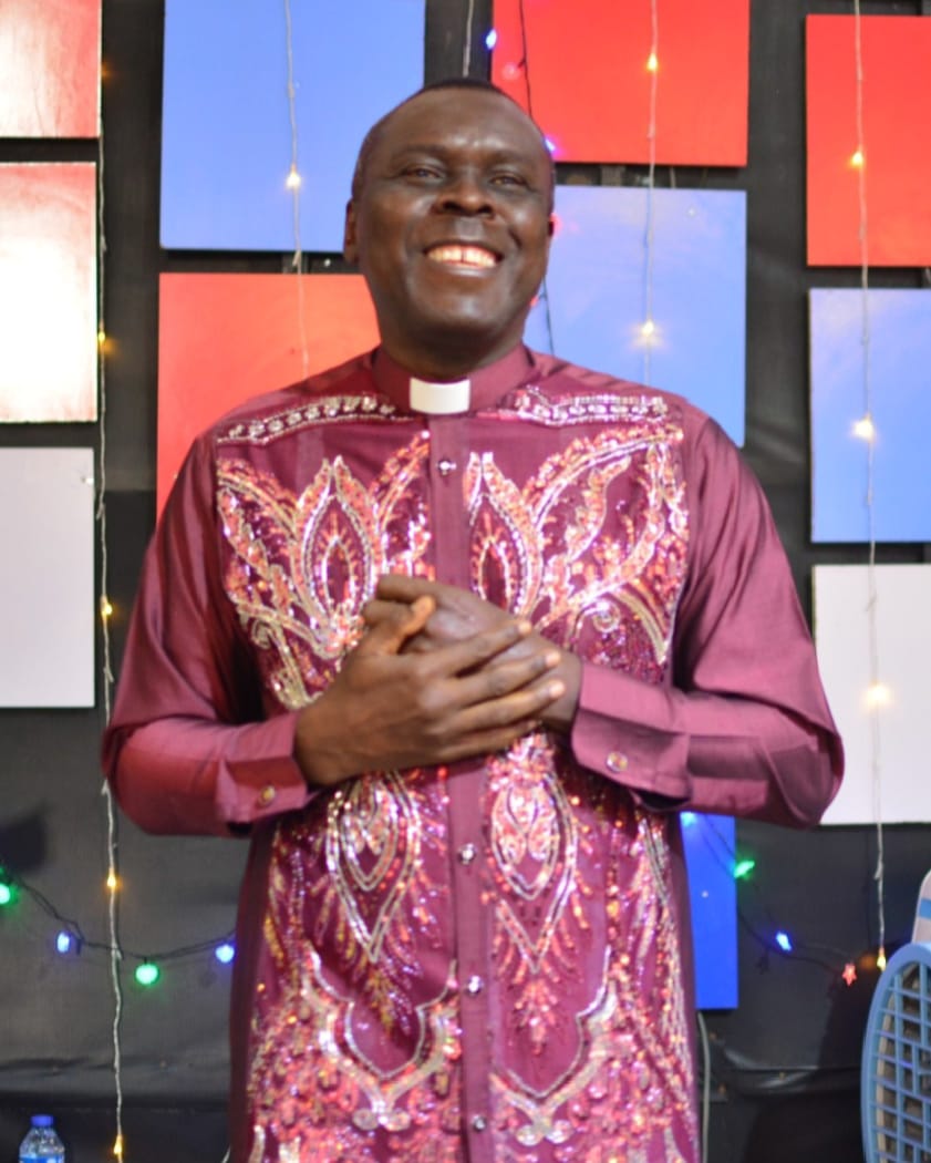 Apostle Prof. E. C. Ejiofor smiling in a maroon clerical shirt with sequined embroidery, hands clasped over his heart