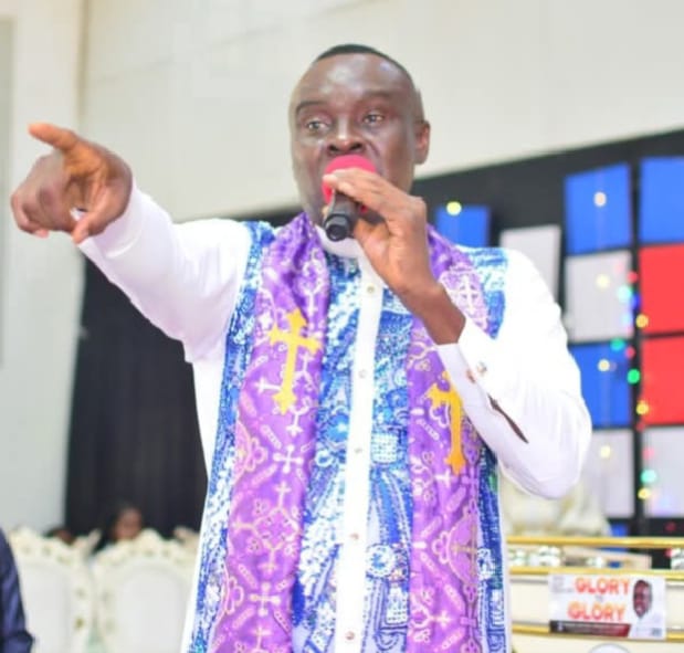 Apostle Prof. E. C. Ejiofor in a patterned clerical stole, speaking into a microphone behind a podium with Glory to Glory inscription