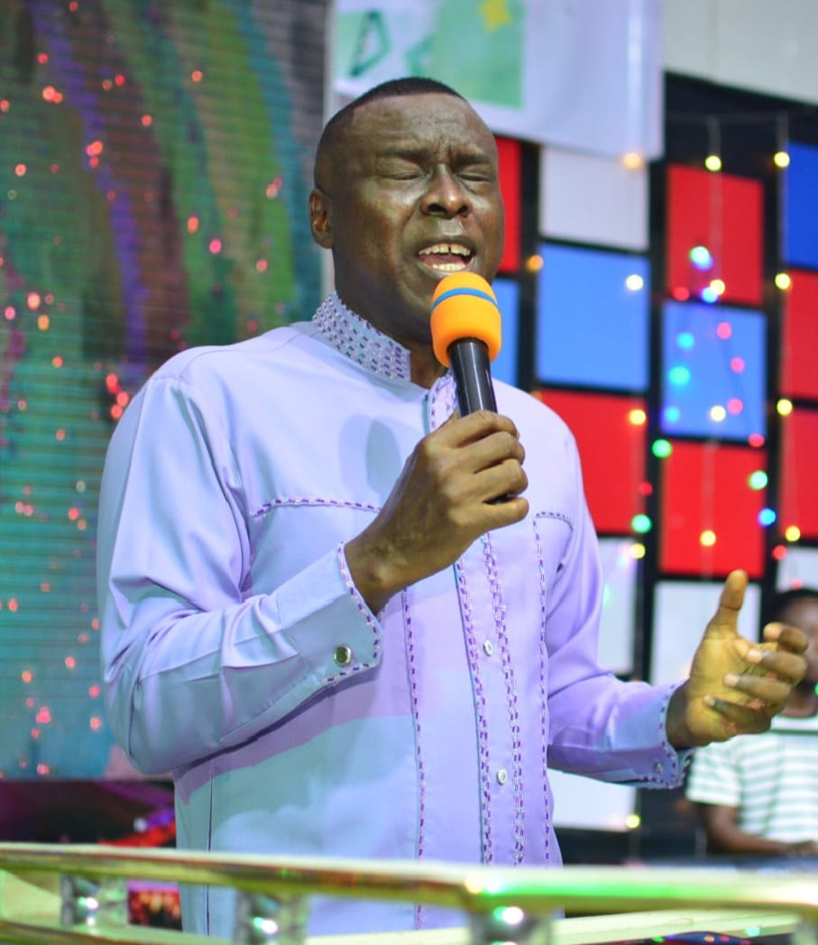 Apostle Prof. E. C. Ejiofor singing into a microphone with an orange windscreen, wearing a lavender tunic with purple and white beaded embroidery — General Overseer of Praiseland Royal Springs Int'l Church