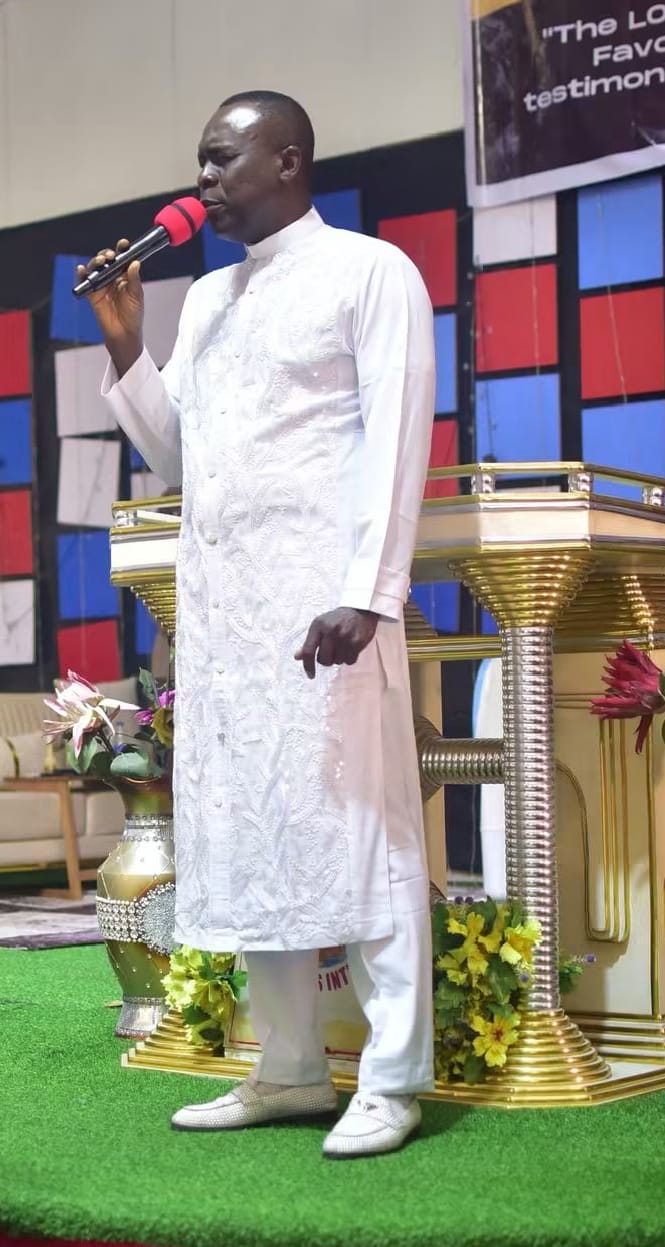 Apostle Prof. E. C. Ejiofor in a white embroidered robe, ministering with a red microphone beside the pulpit