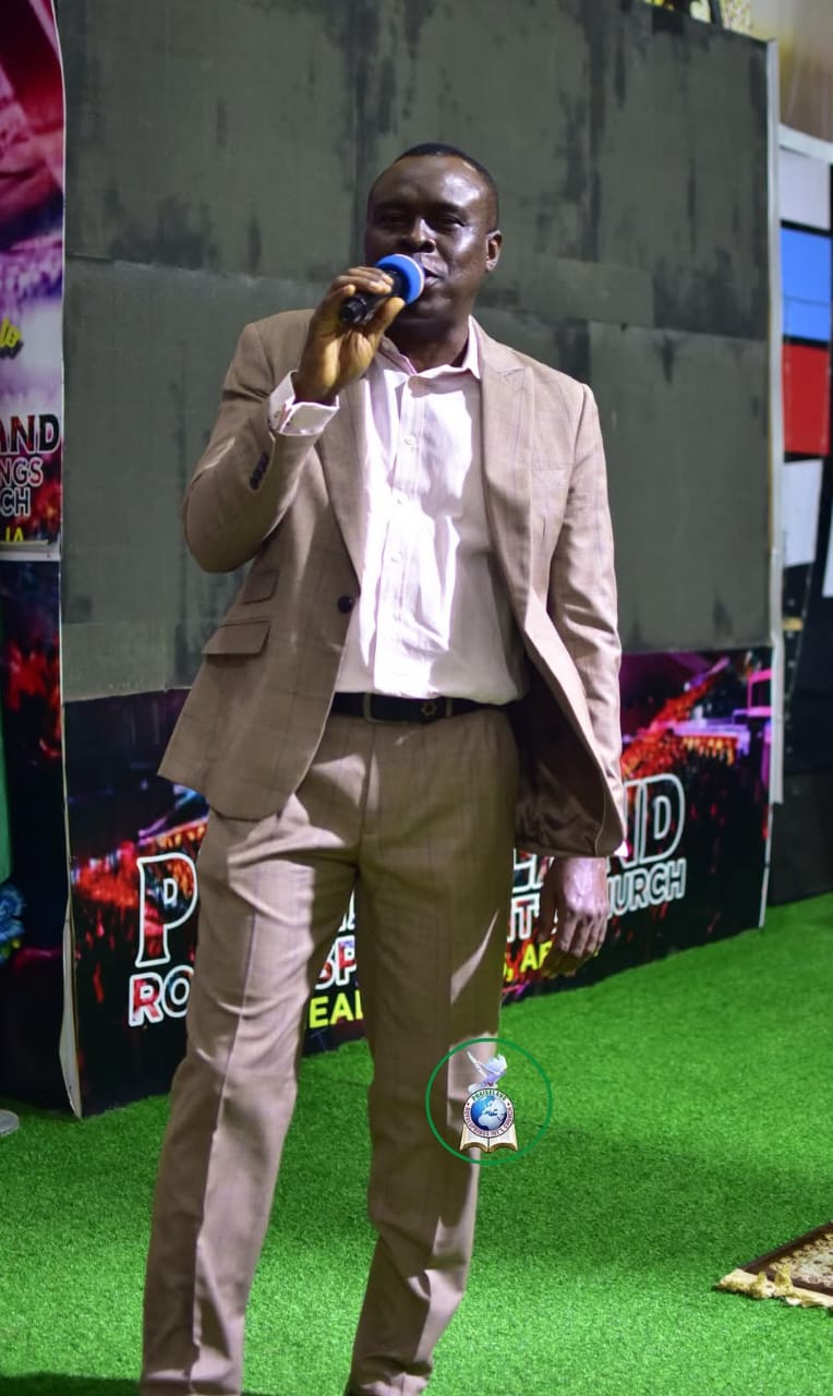 Apostle Prof. E. C. Ejiofor on stage holding a microphone, wearing a light brown suit jacket over a pinkish-white shirt — General Overseer of Praiseland Royal Springs Int'l Church