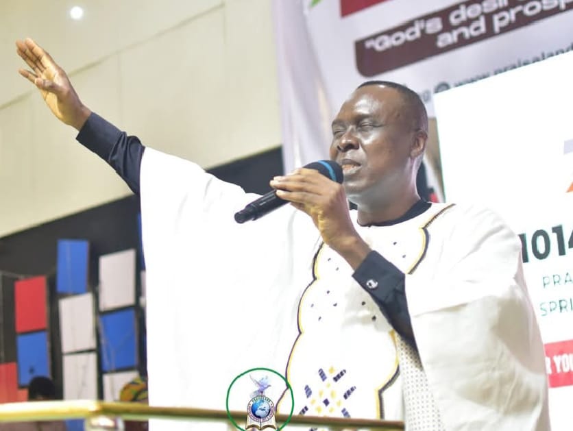Apostle Prof. E. C. Ejiofor ministering with a raised hand, wearing a white embroidered robe