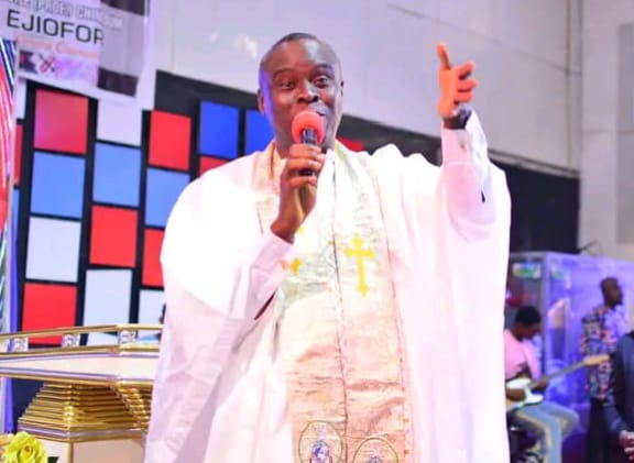 Apostle Prof. E. C. Ejiofor preaching into a microphone with his hand raised, wearing a white liturgical robe and a white stole embroidered with gold crosses, framed by a colourful red, blue and white panelled wall