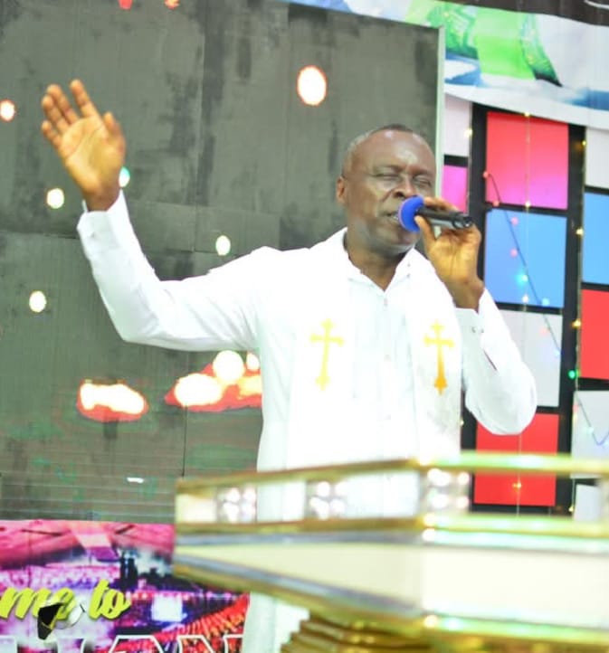 Apostle Prof. E. C. Ejiofor preaching into a microphone with his left arm raised and palm open, wearing a white garment with a white stole featuring gold crosses, on a church stage with colored panel backdrop