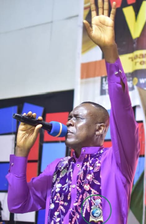 Apostle Prof. E. C. Ejiofor singing into a microphone with his left arm raised and open palm, wearing a bright purple shirt with an embroidered white and light purple front panel, in a studio with a wall of blue, red and white panels behind him