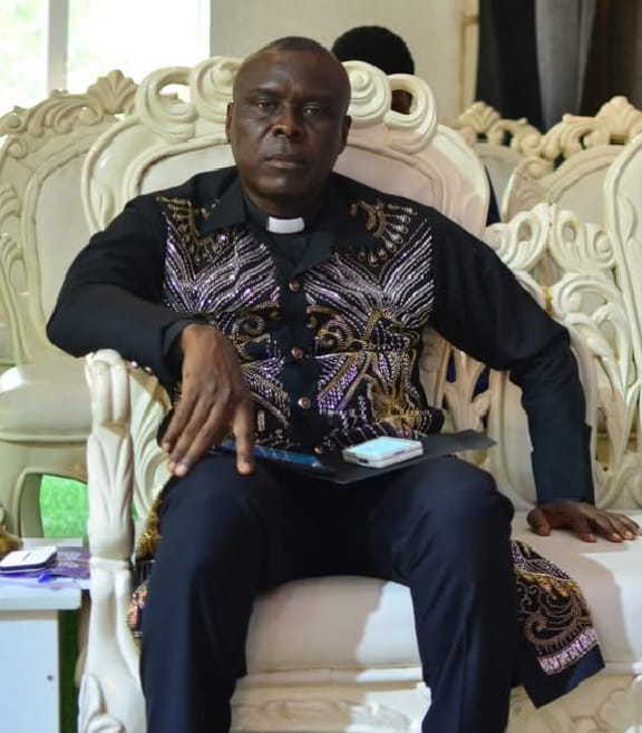 Apostle Prof. E. C. Ejiofor seated in an ornate chair in a place of worship, wearing a black clerical shirt with silver and gold embroidered chest panel and white tab collar — General Overseer of Praiseland Royal Springs Int'l Church