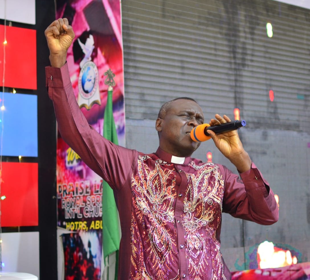 Apostle Prof. E. C. Ejiofor preaching in a maroon embroidered robe, holding a microphone with one fist raised at Praiseland Royal Springs Int'l Church