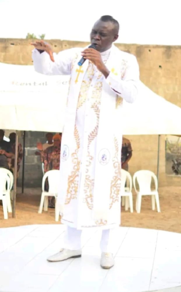 Apostle Prof. E. C. Ejiofor ministering in a white and gold robe with a microphone at an open-air service