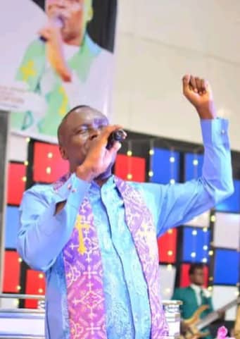 Apostle Prof. E. C. Ejiofor ministering with a microphone, hand raised in worship, wearing a blue and purple embroidered outfit