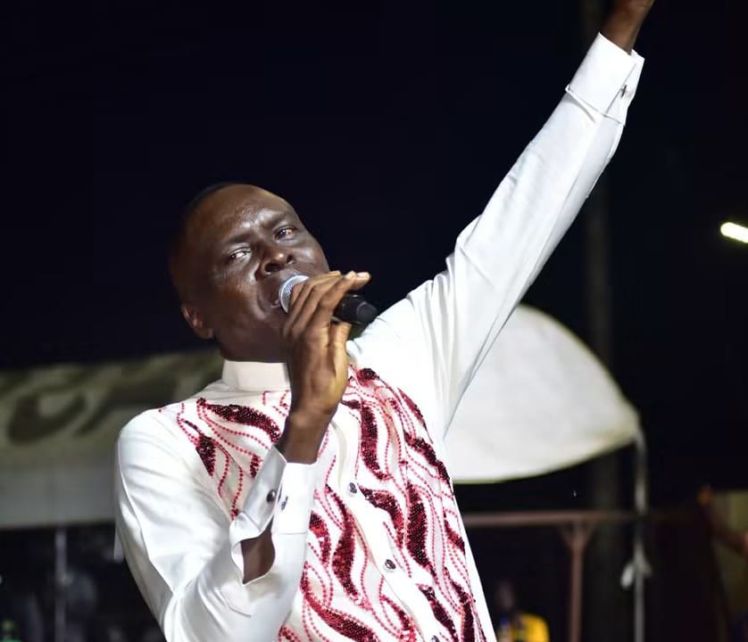 Apostle Prof. E. C. Ejiofor preaching with a microphone and one arm raised, wearing a white shirt with flame-like red embroidery