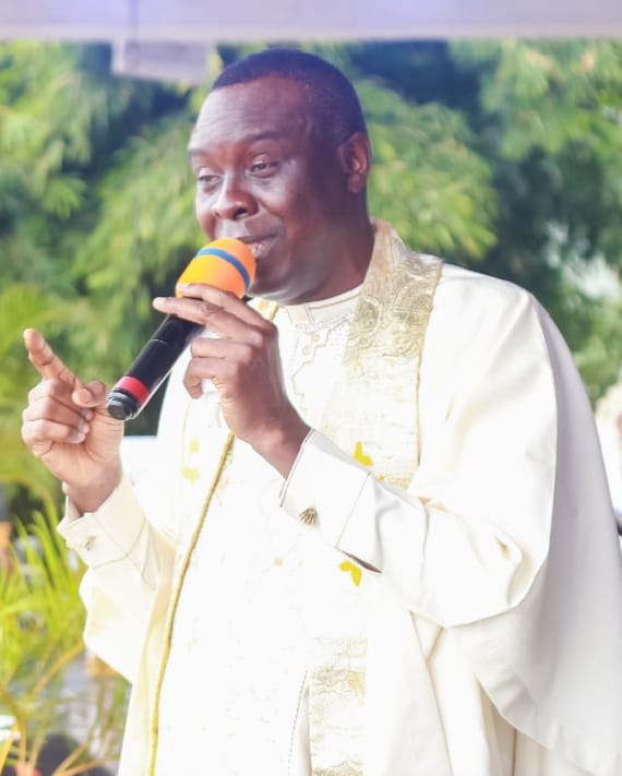 Apostle Prof. E. C. Ejiofor preaching outdoors with a microphone, wearing an off-white clerical robe with a gold-embroidered cream stole and white tab collar, pointing upward — General Overseer of Praiseland Royal Springs Int'l Church