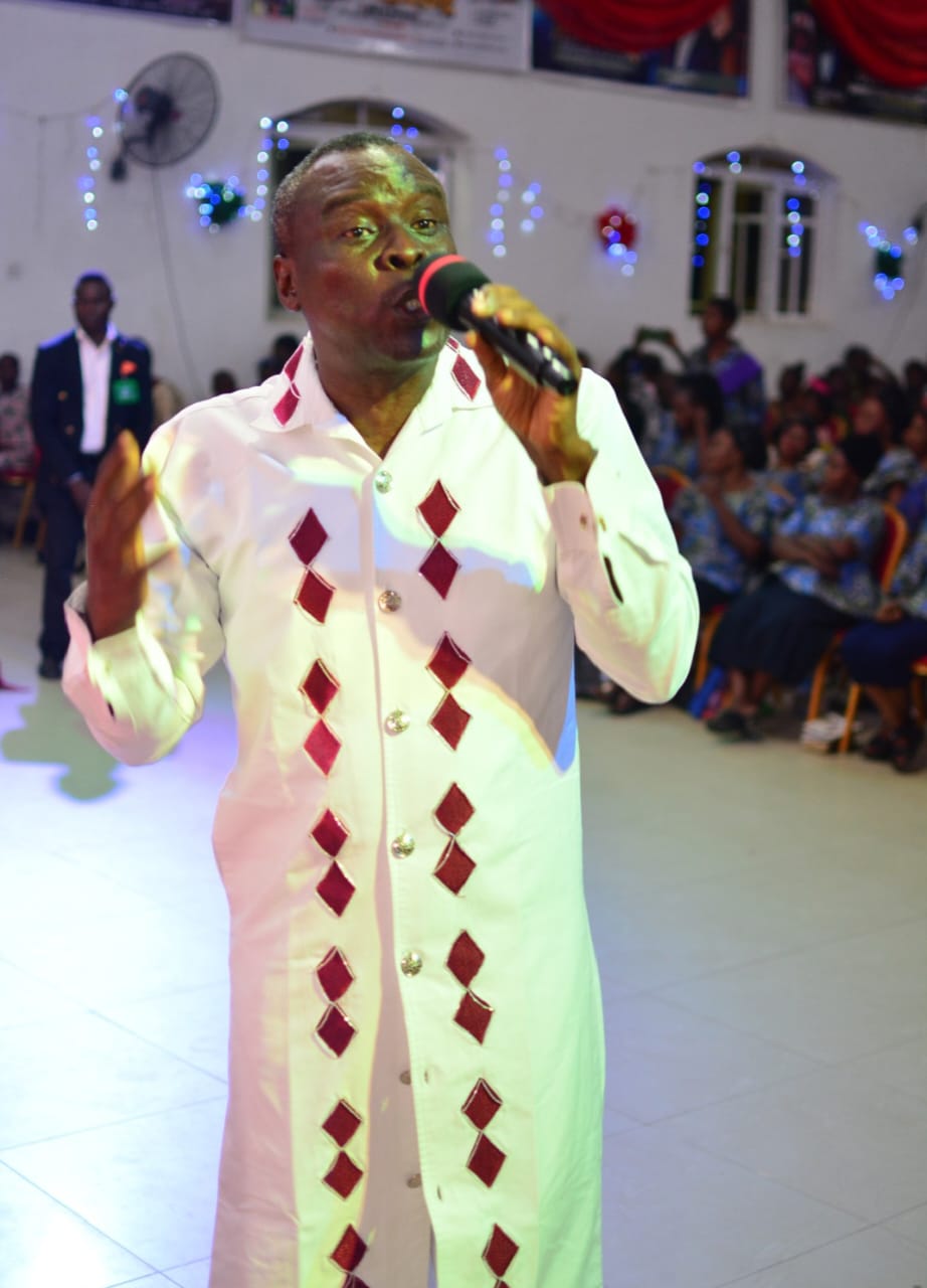 Apostle Prof. E. C. Ejiofor preaching into a microphone, wearing a white tunic with red diamond embroidery and silver buttons