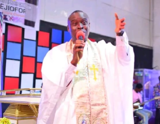Apostle Prof. E. C. Ejiofor preaching with a microphone, one arm raised, wearing a white robe with a gold-embroidered stole