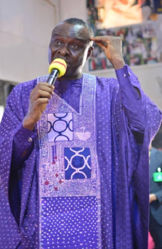 Apostle Prof. E. C. Ejiofor preaching into a microphone with his hand raised toward his forehead, wearing a vibrant purple Agbada robe with intricate lavender and blue-purple embroidery panels