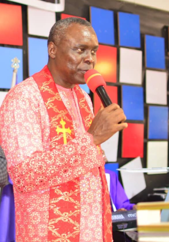 Apostle Prof. E. C. Ejiofor preaching with a microphone, wearing a red patterned robe with a gold cross stole