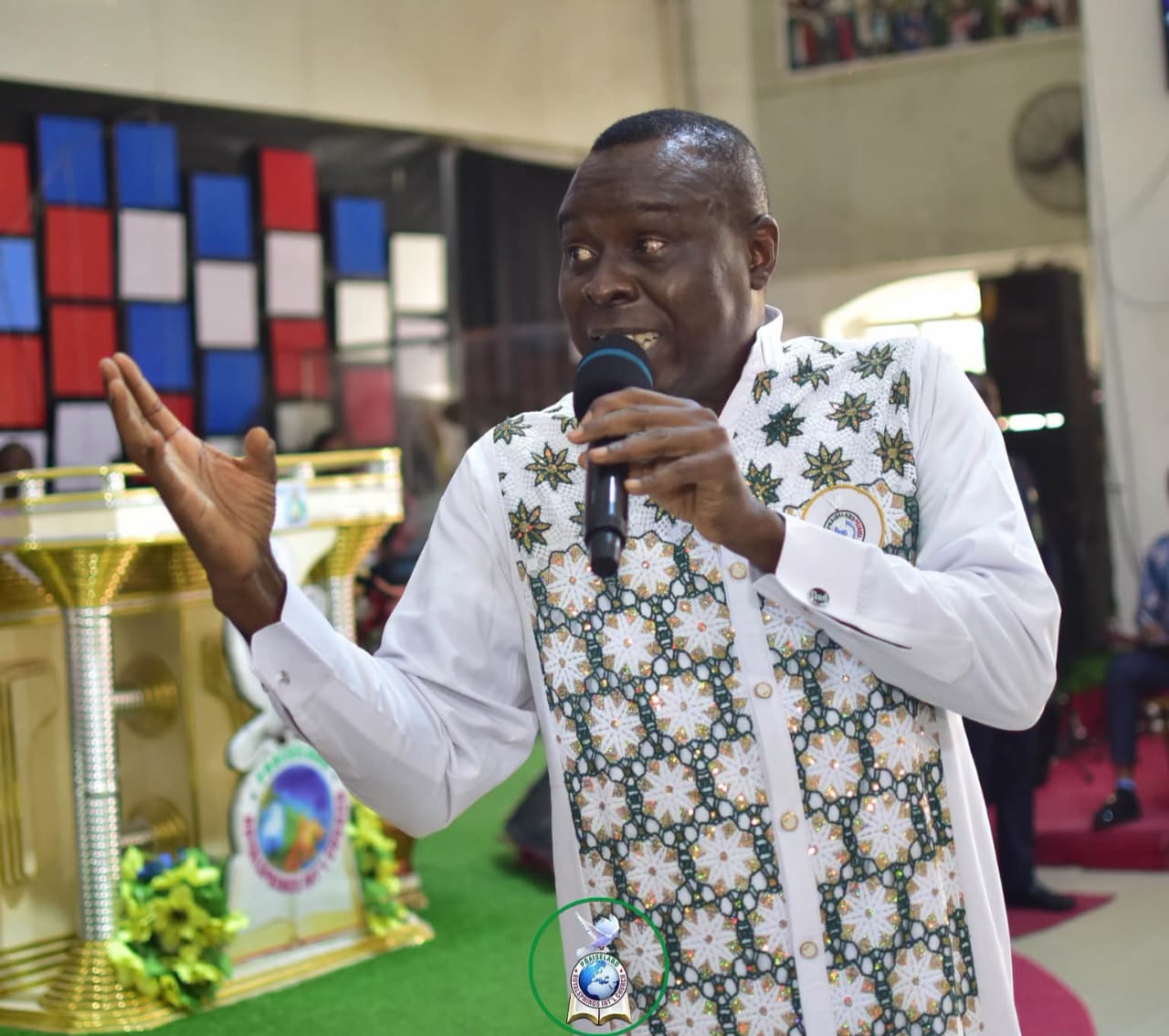 Apostle Prof. E. C. Ejiofor preaching with a microphone, wearing a white and green embroidered outfit
