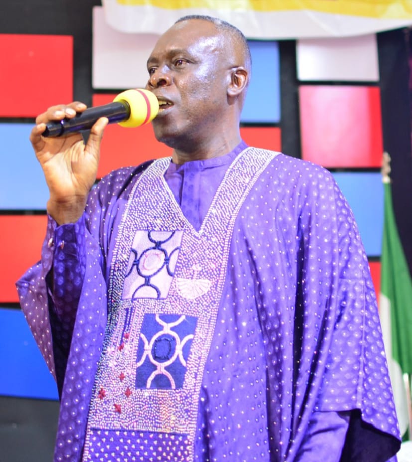 Apostle Prof. E. C. Ejiofor in a purple agbada with silver beaded embroidery, singing into a microphone with a yellow windscreen on stage — General Overseer of Praiseland Royal Springs Int'l Church