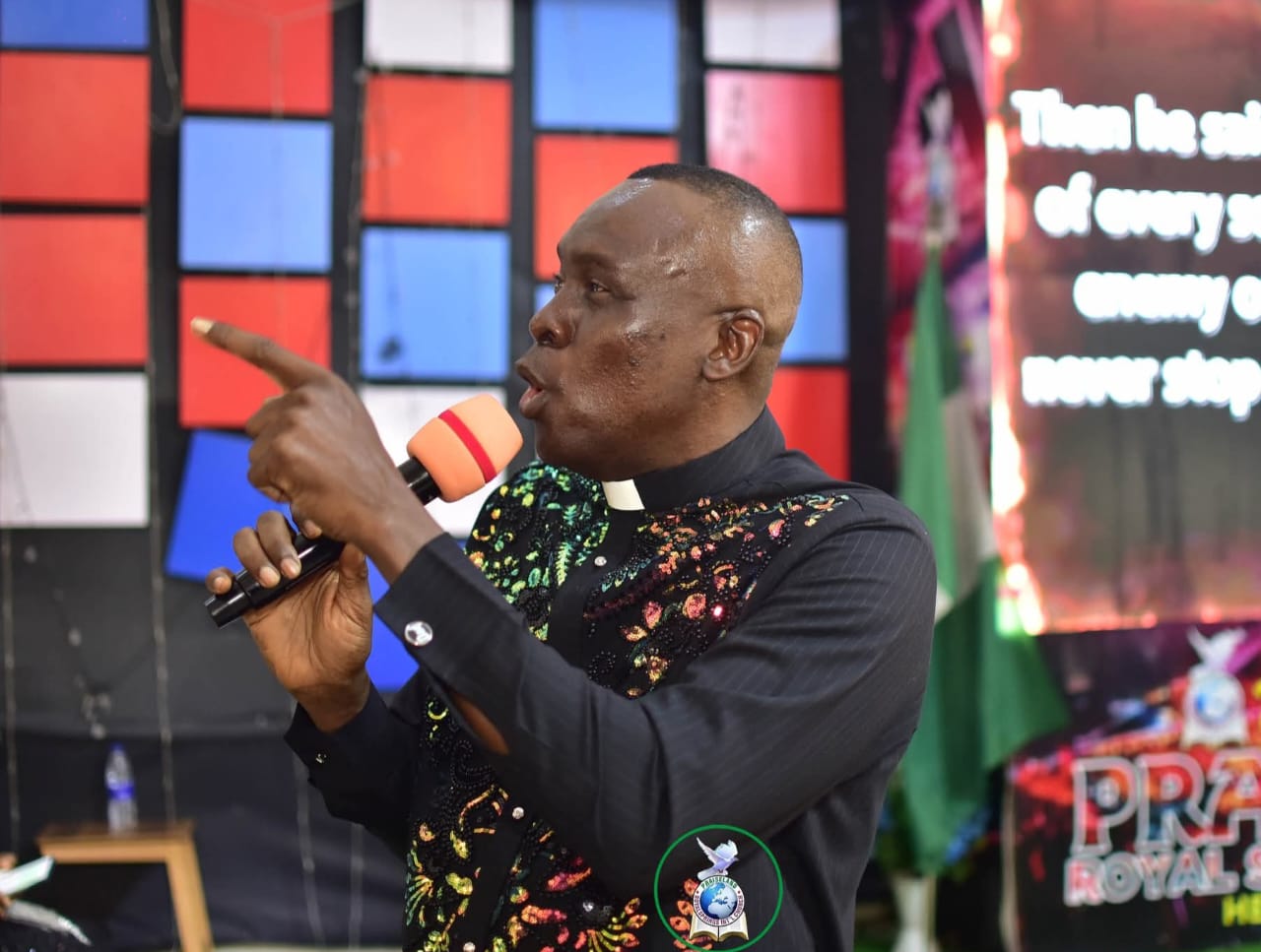 Apostle Prof. E. C. Ejiofor in a black clerical robe with a white collar and shimmering multi-coloured sequin embroidery, speaking into a microphone with an orange windscreen and pointing forward with his right hand, on a church stage with a Nigerian flag and Praiseland Royal Springs banner behind him