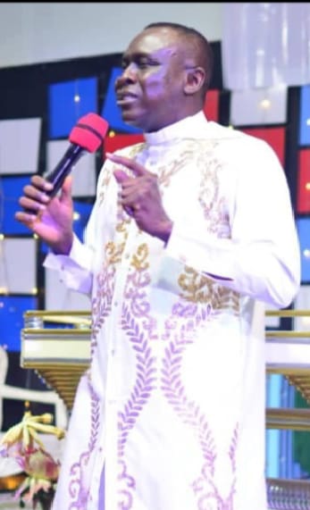 Apostle Prof. E. C. Ejiofor preaching with a microphone, one hand raised with index finger pointing upward, wearing a white robe with gold embroidery
