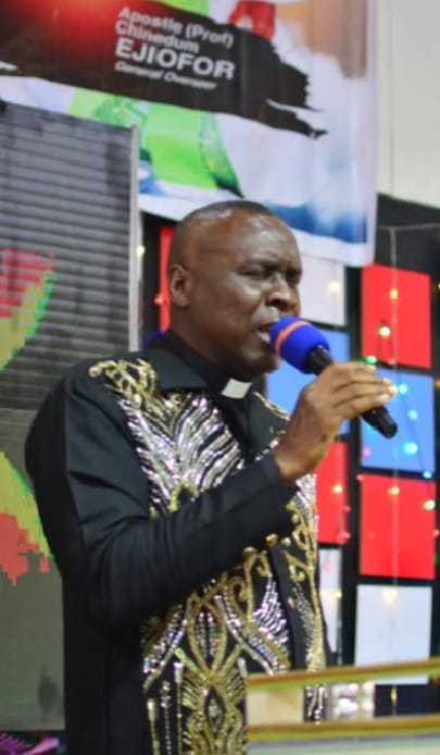 Apostle Prof. E. C. Ejiofor preaching with a microphone, wearing a black robe with gold and silver embroidery
