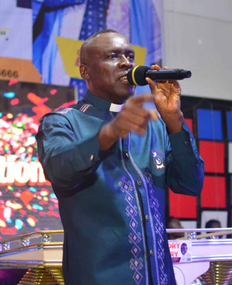 Apostle Prof. E. C. Ejiofor preaching into a microphone, wearing a dark teal-blue clerical shirt with a white collar and a purple-and-white diamond-patterned decorative band, pointing toward the congregation